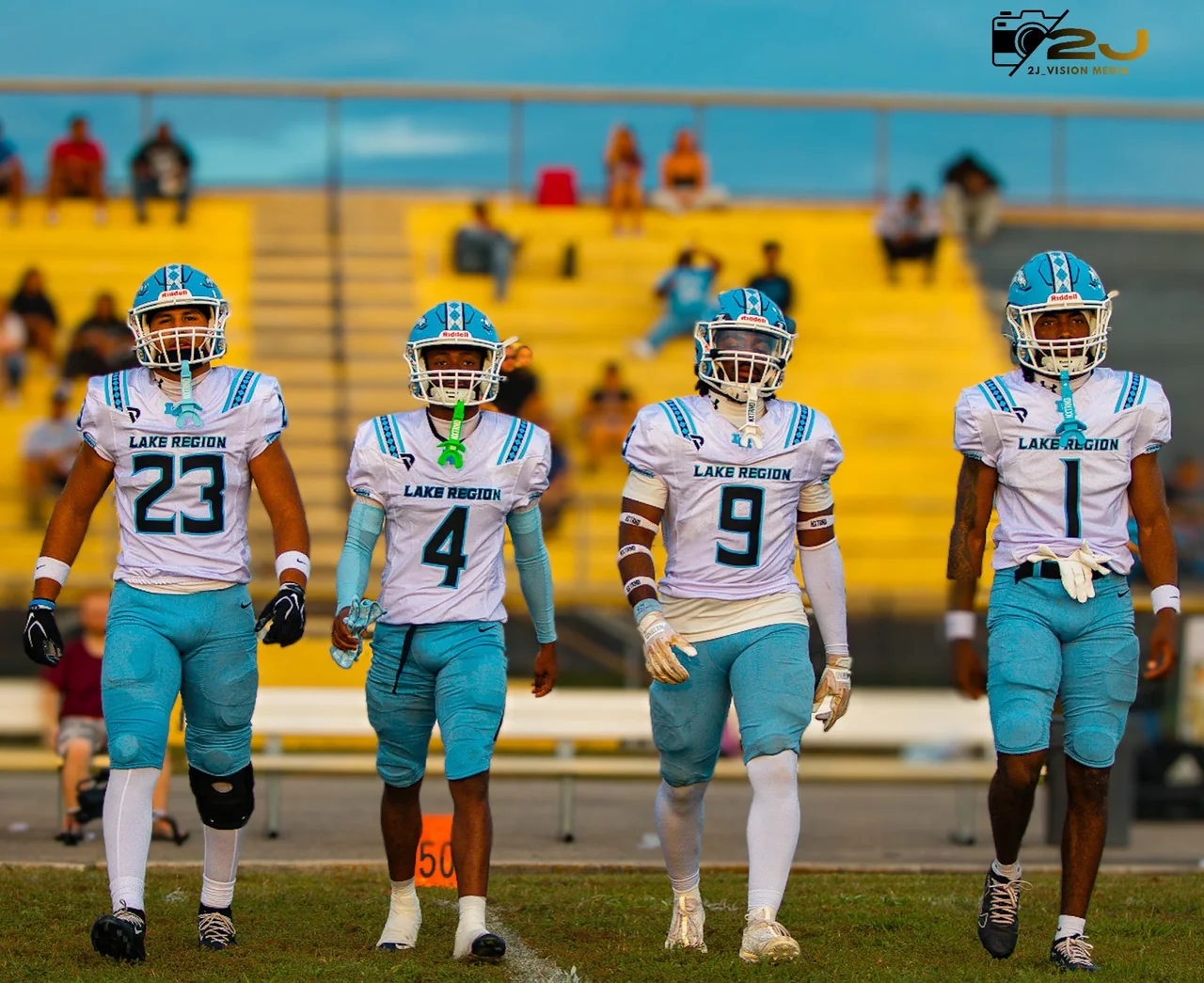 Four Lake Region football players walking together in white jerseys and turquoise pants, with numbers 23, 4, 9 and 1; original 2J Vision Media watermark visible